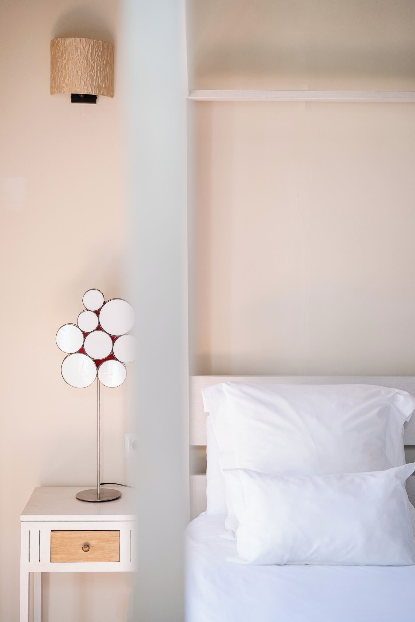 A neatly arranged bedroom with a white bed, a bedside table with a lamp, and a wall-mounted light fixture.