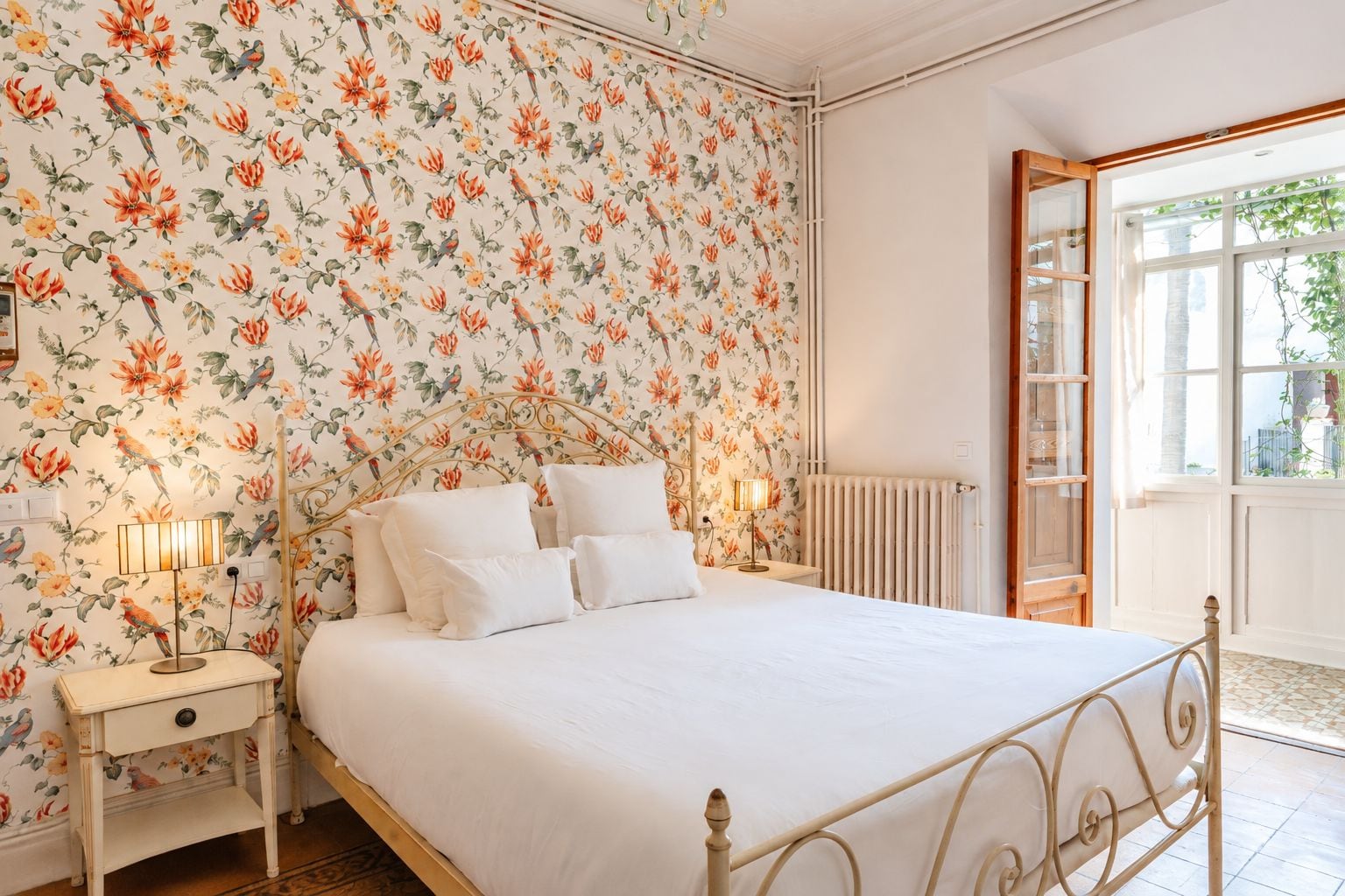 A bedroom with a floral wallpaper, a bed with white bedding, two bedside lamps, and a window with a view of greenery.