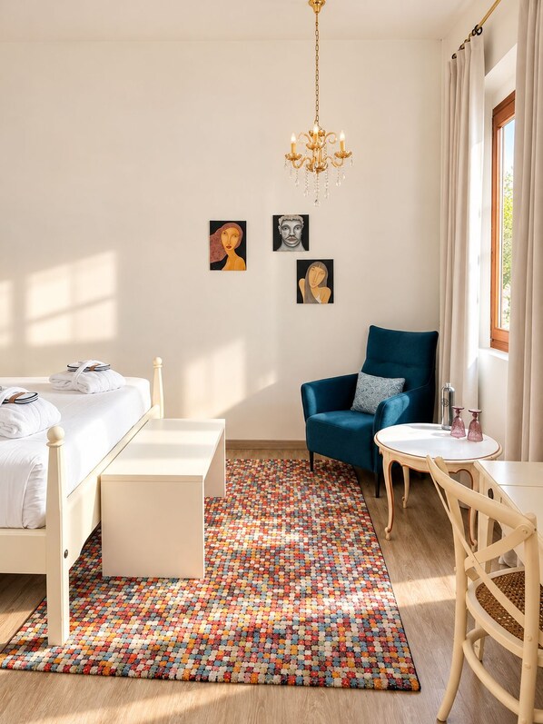 A hotel room with a bed, a blue armchair, a small table, and a colorful rug. There are three paintings on the wall. - Hotel Can Abril (Sóller)