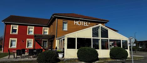 A two-story hotel with a red and brown facade, a covered outdoor seating area, and a sign indicating the establishment is a hotel.