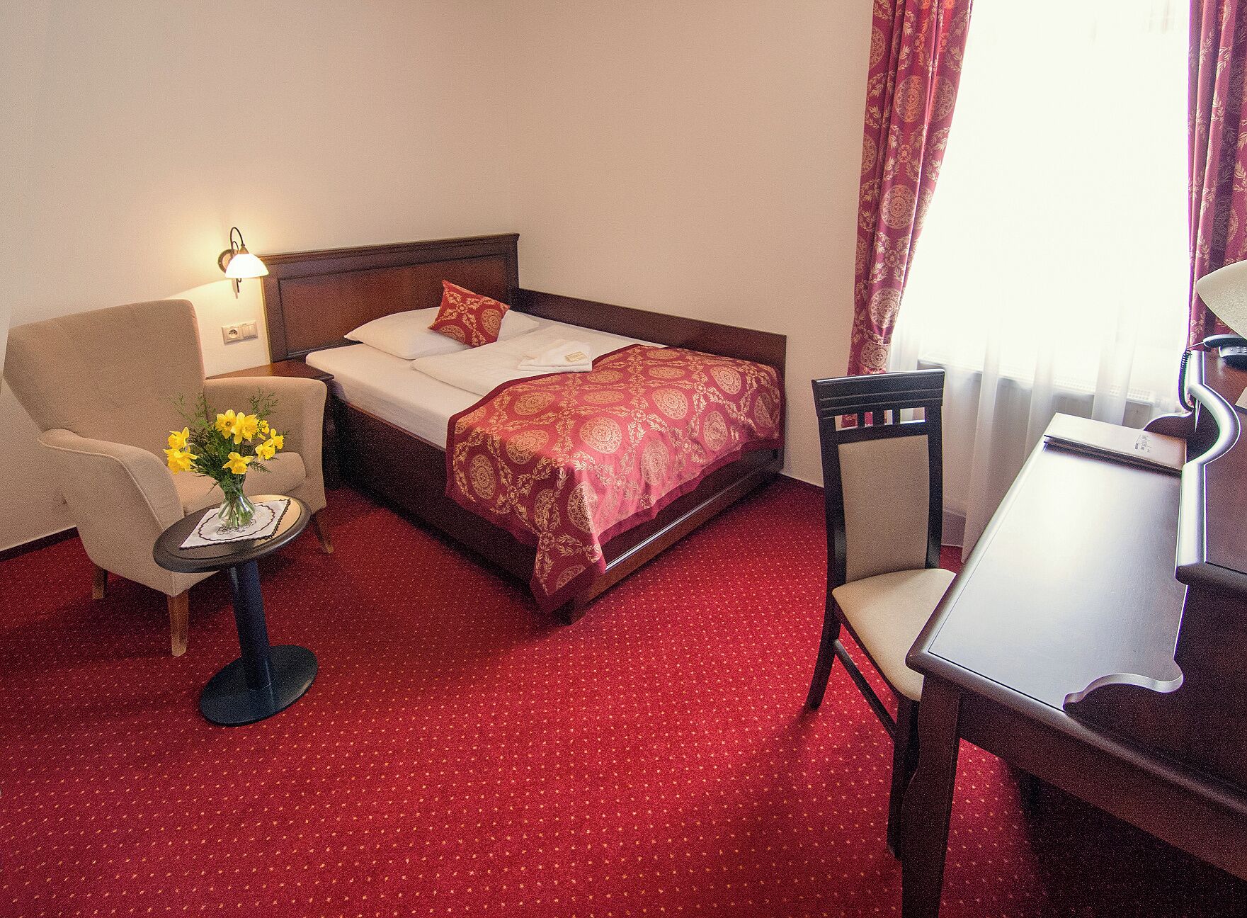 Una habitación de hotel con cama, una silla, una mesita con un jarrón de flores y un escritorio con lámpara.