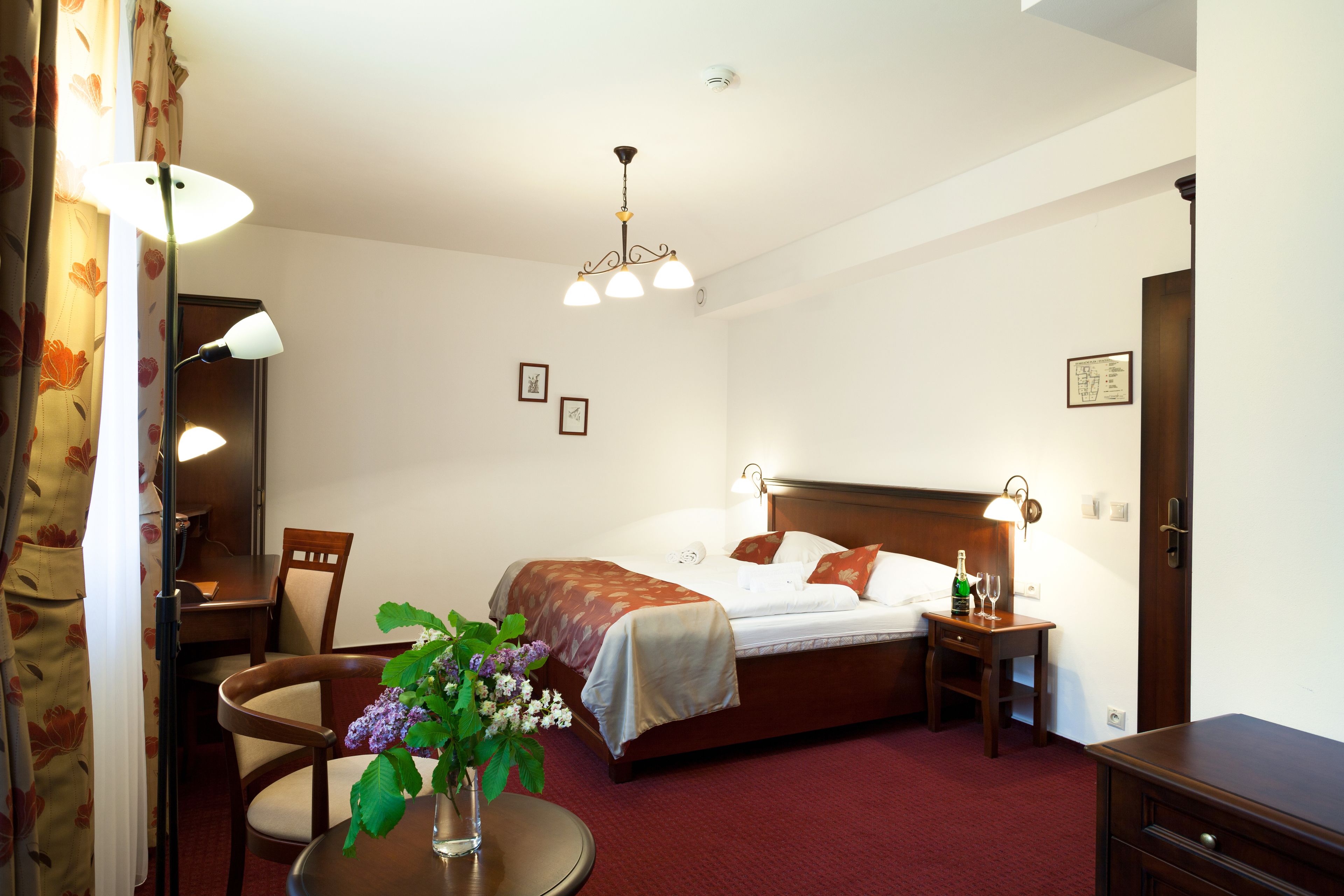 A hotel room with a bed, bedside table, lamp, chair, and a small table with a vase of flowers.