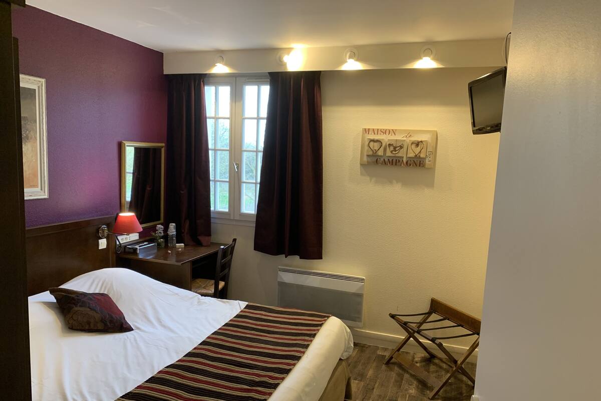 A hotel room with a bed, a desk, a chair, a radiator, and a television mounted on the wall.