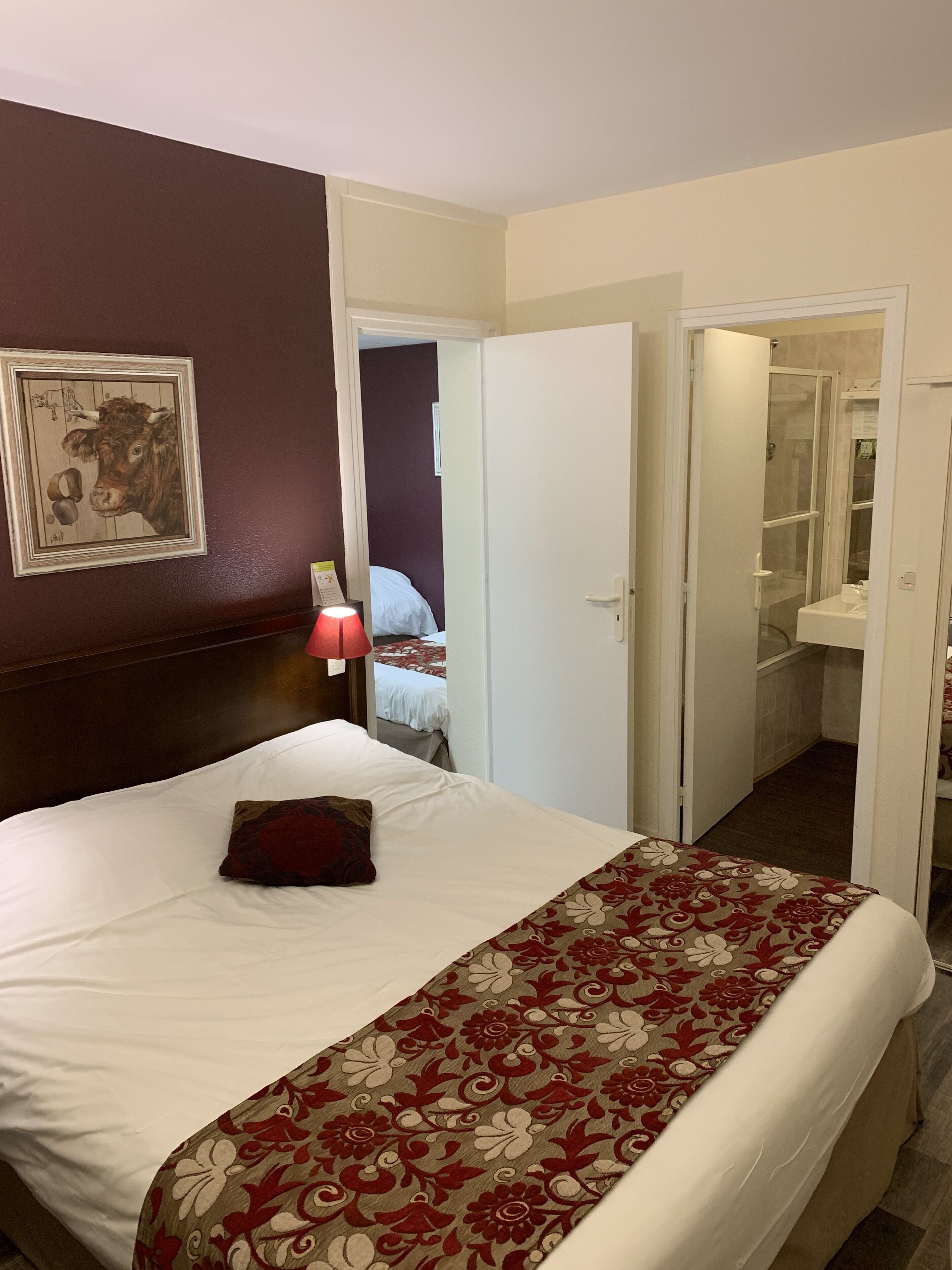 A hotel room with a large bed, a nightstand with a lamp, a framed picture on the wall, and a bathroom visible through an open door.