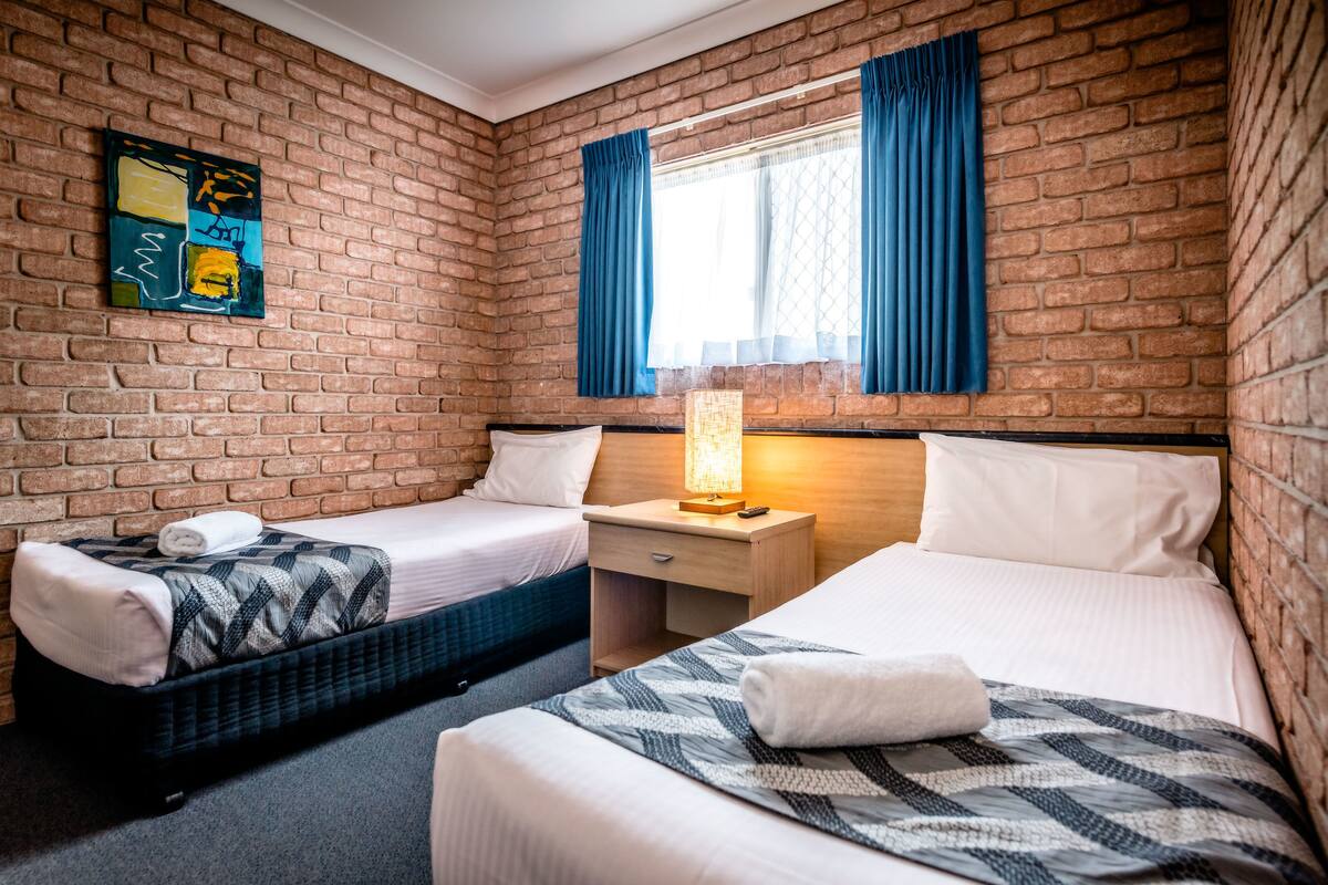 A hotel room with a brick wall, two beds, a nightstand, a lamp, and a window with blue curtains.