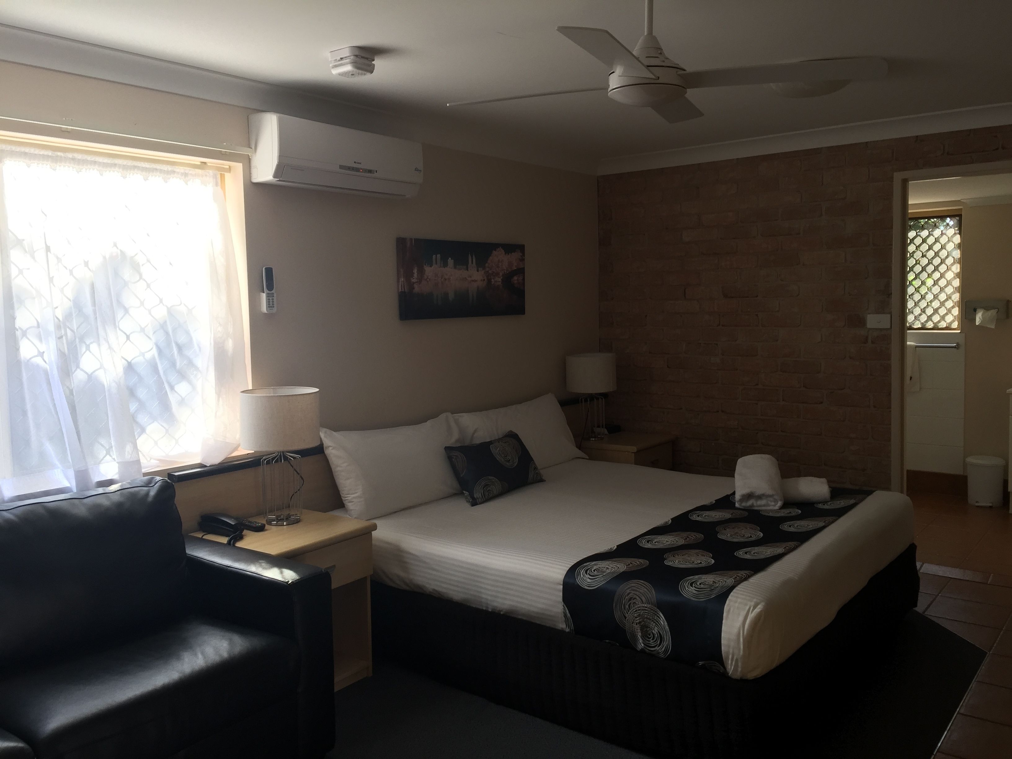 A hotel room with a bed, a desk with a lamp, a chair, a ceiling fan, and a window with curtains.