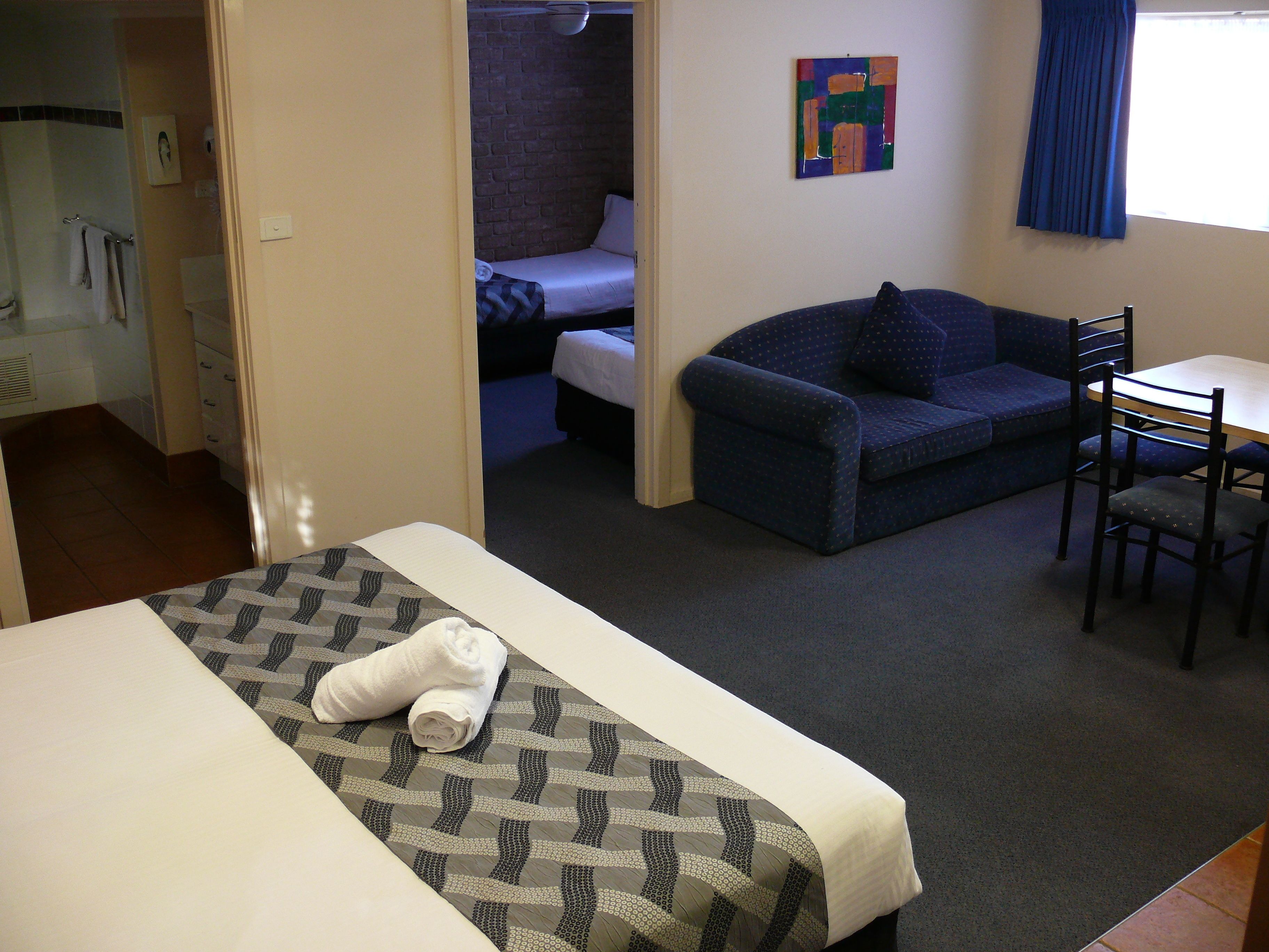 A hotel room with a bed, a sofa, a dining table with chairs, a bathroom, and a window with curtains.
