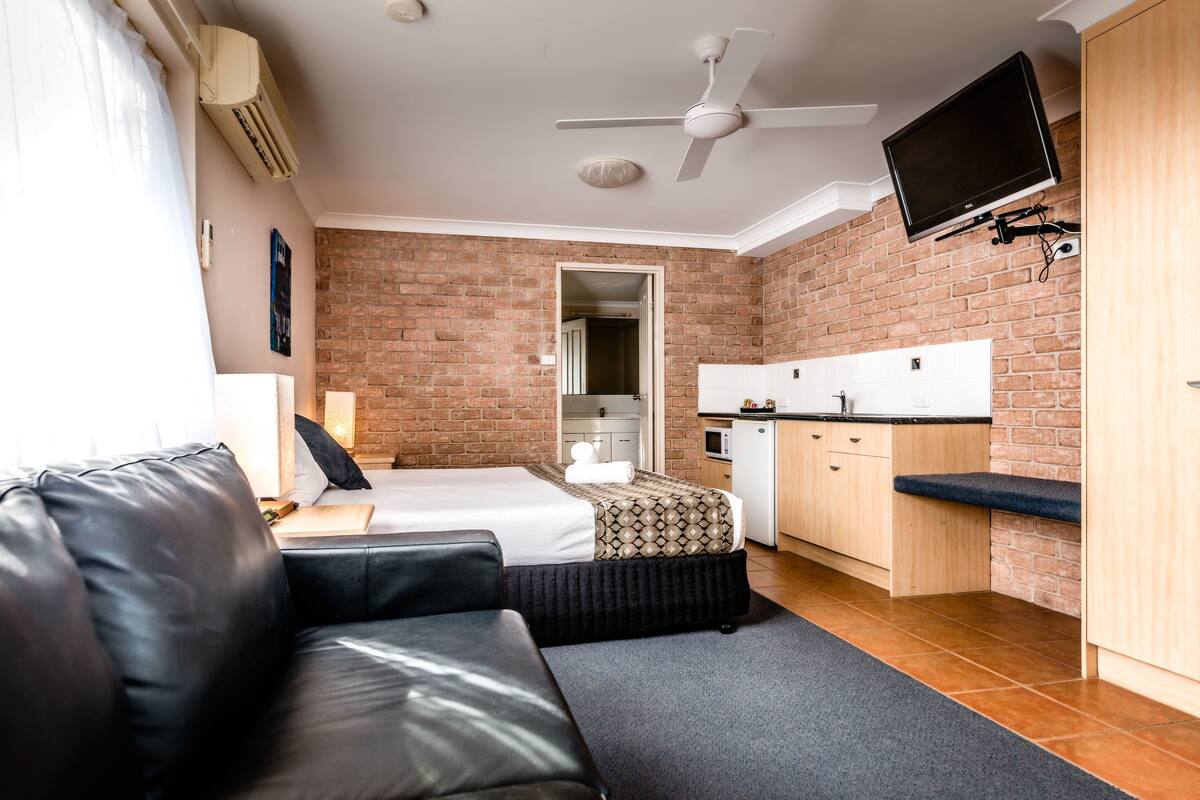A compact room with a bed, a sofa, a small kitchenette, and a TV mounted on the wall.