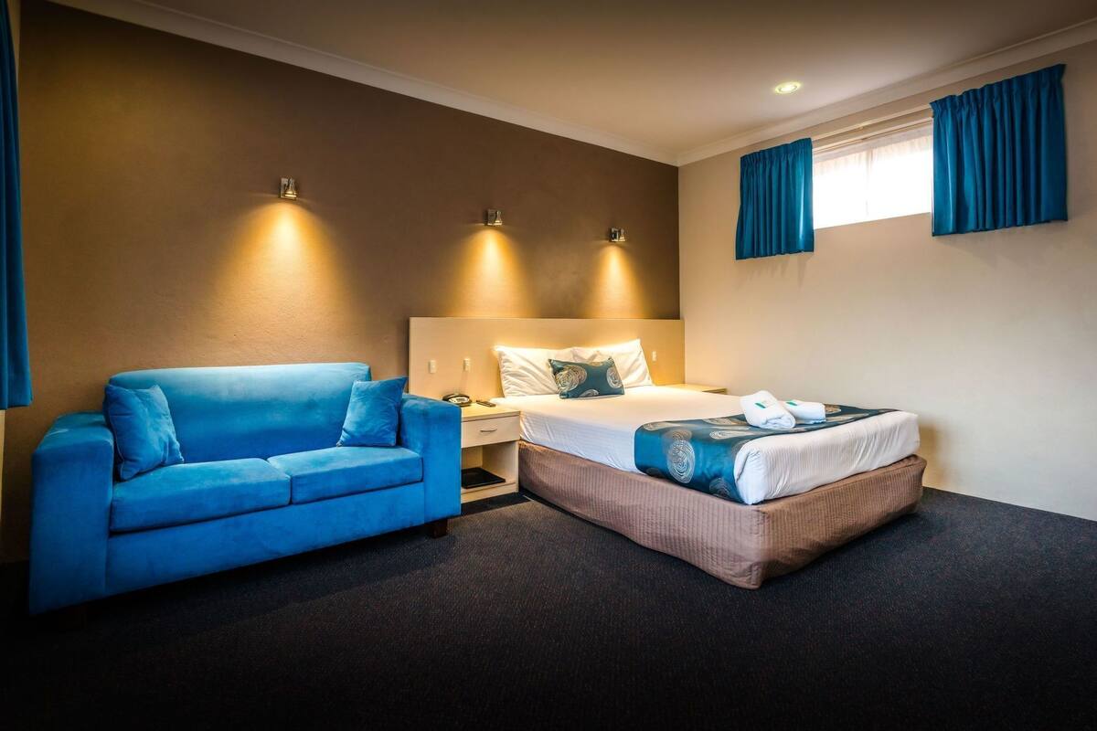 A hotel room with a blue sofa, a bed with a patterned blanket, and two wall-mounted lights.