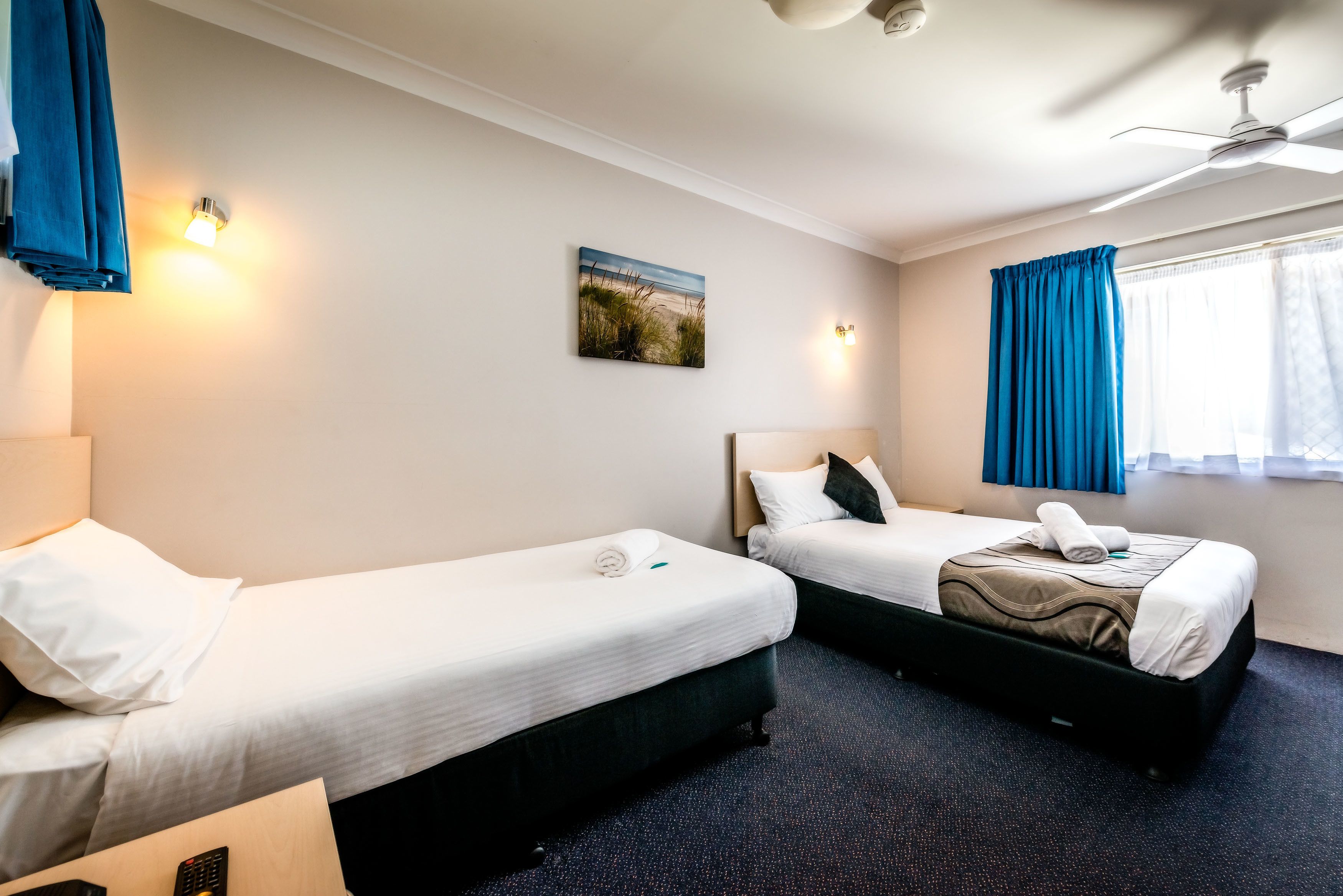 A hotel room with two beds, a ceiling fan, a painting, and a window with curtains.
