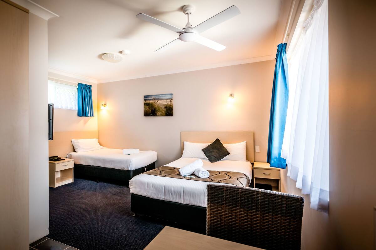 A hotel room with two beds, a ceiling fan, a painting on the wall, and a window with curtains.