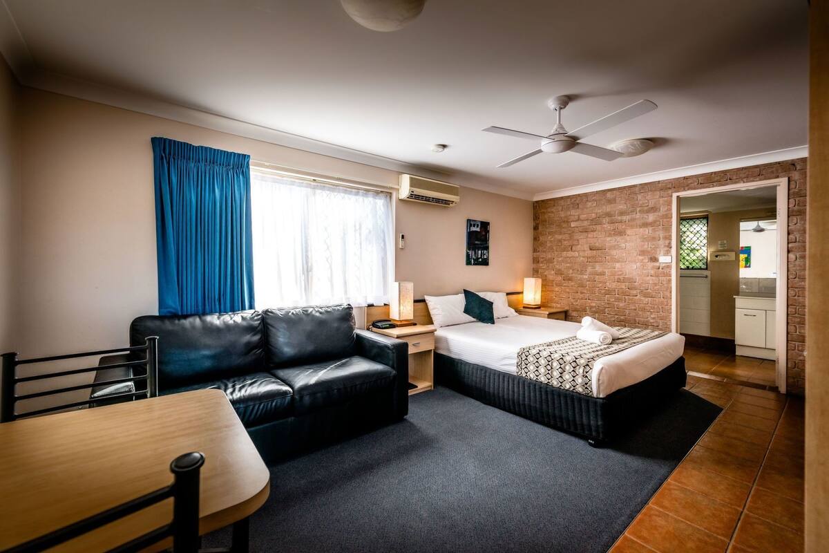A hotel room with a bed, a sofa, a desk, a chair, a ceiling fan, and a brick wall.