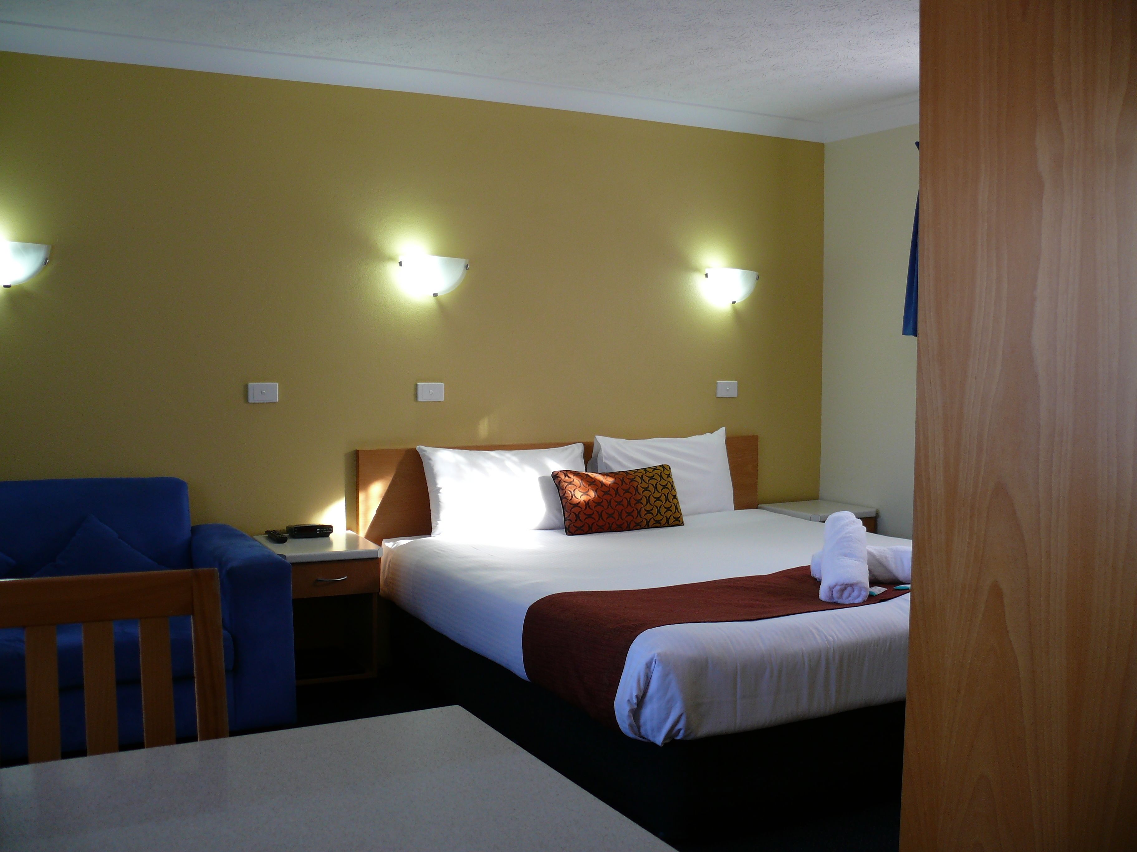 A hotel room with a bed, a desk, and a chair. There are two wall-mounted lights and a door.