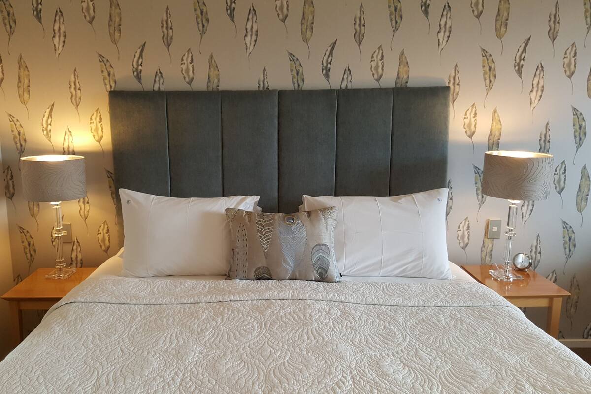 A neatly made bed with a patterned bedspread, a dark upholstered headboard, and two bedside lamps.