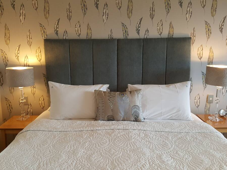 Een net opgemaakt bed met een beddegoed met een patroon, een donker bekleed hoofdeinde en twee bedlampjes.