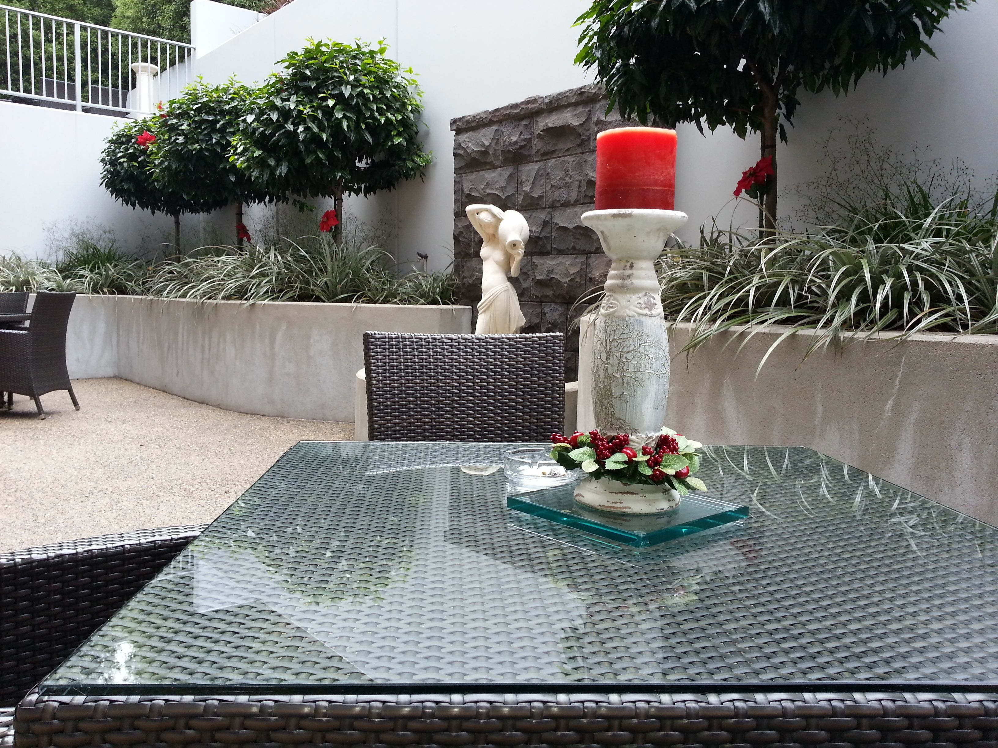 Uma mesa com tampo de vidro, um vaso branco e berras vermelhas, rodeada por cadeiras de vime e uma pequena estátua.