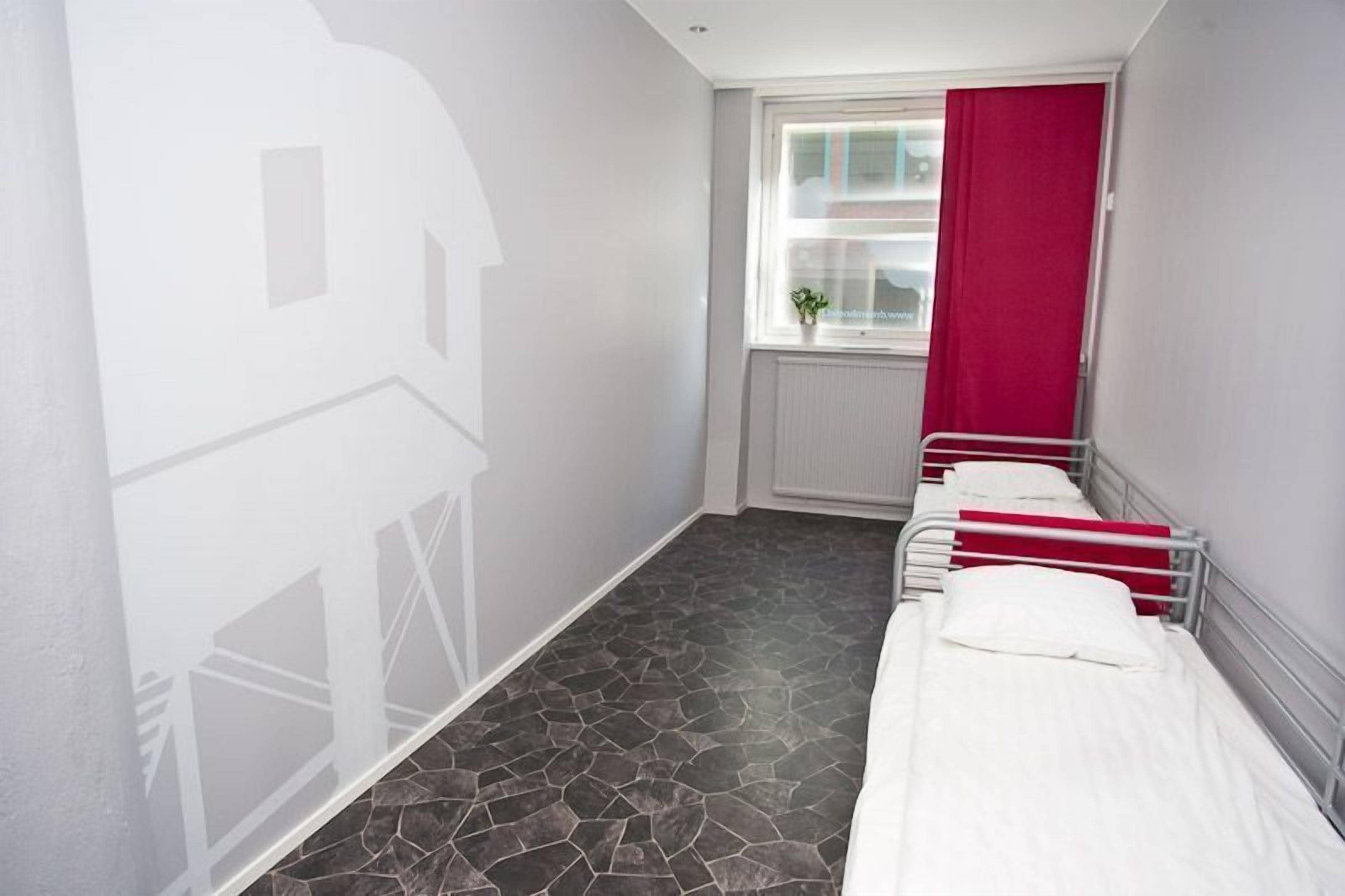 Quarto de hotel com três camas, uma janela com cortina vermelha e piso com padrão de pedra.