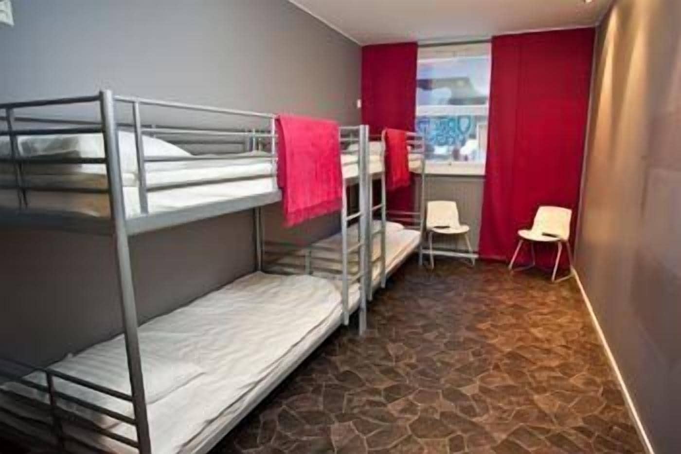 Una habitació de dormitori amb lliteres, una taula petita, dues cadires i una finestra amb cortines vermelles.