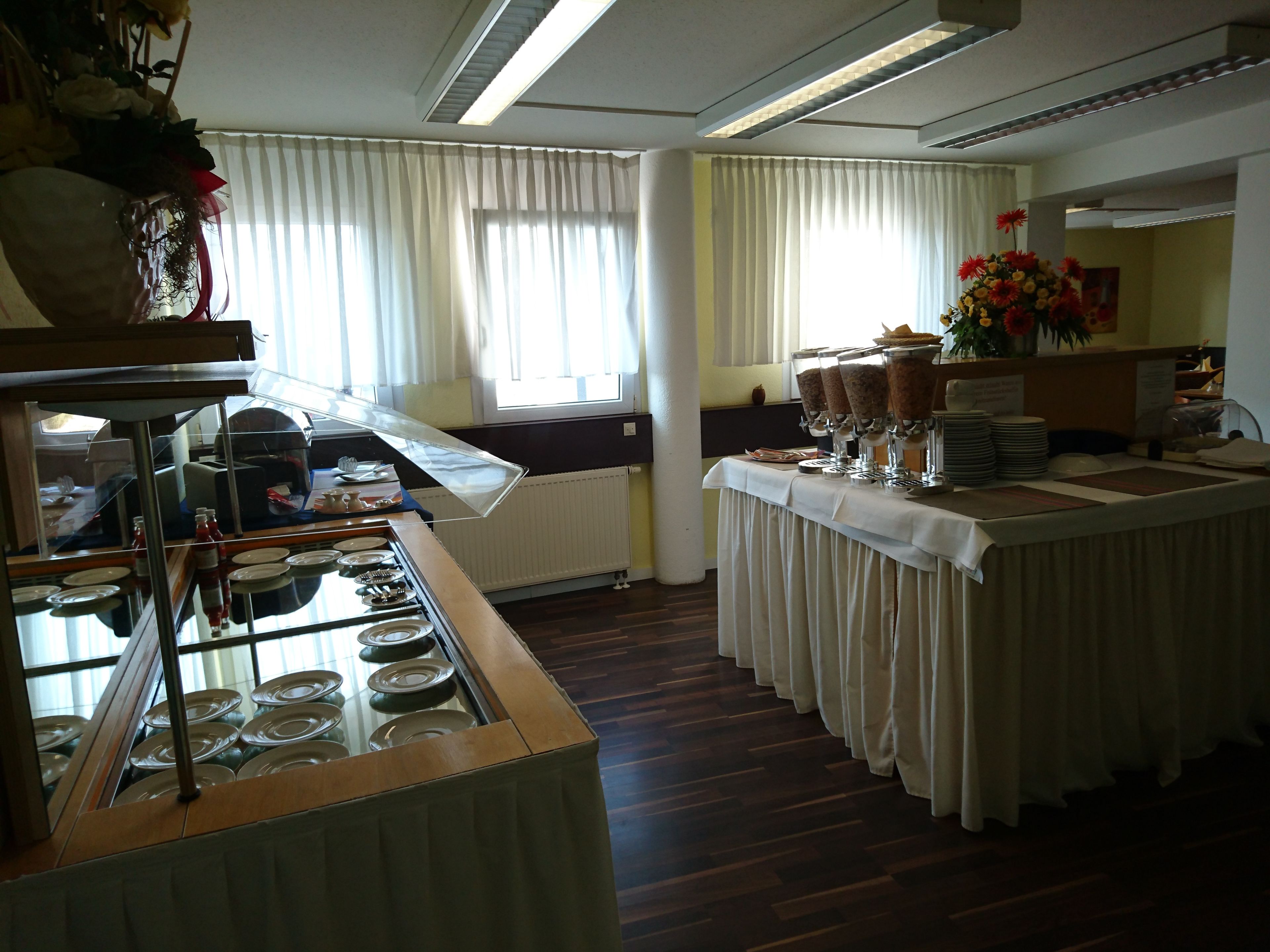 A buffet setup with a variety of food items and a beverage station, a floral arrangement, and a window with curtains.