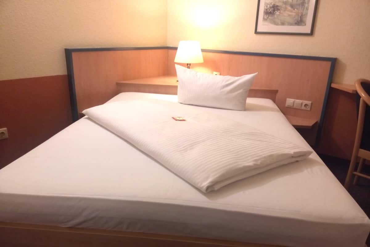 A neatly made bed with white linens, a single pillow, and a bedside lamp.