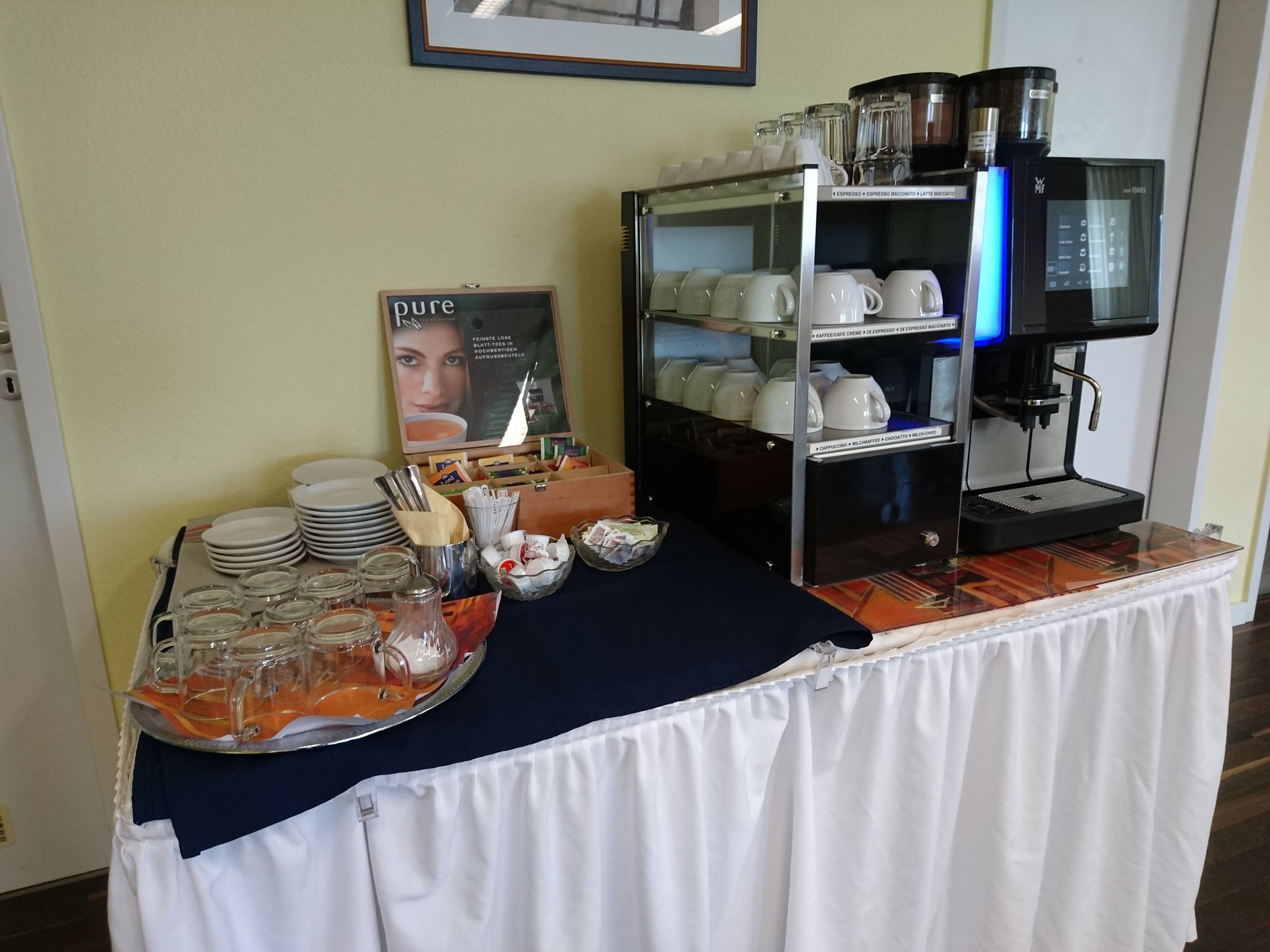 A buffet setup with a coffee machine, plates, glasses, and a sign.