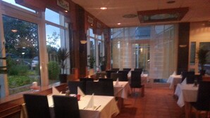 A restaurant with multiple tables and chairs, large windows, and indoor plants. - Central Hotel (Winnenden)