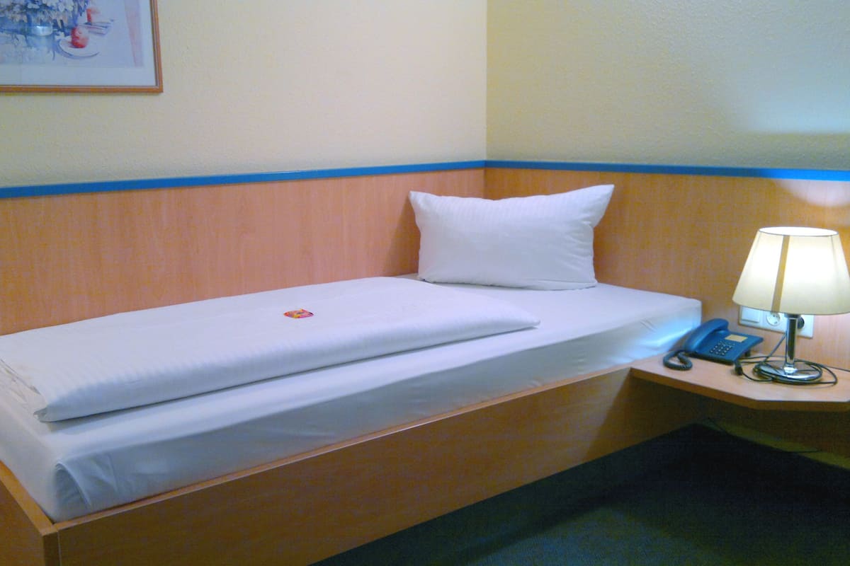 A single bed with a white mattress and pillow, a bedside table with a lamp and a phone, and a framed picture on the wall.