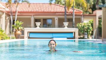A swimming pool with a person in the water, surrounded by palm trees and a building with a red-tiled roof.