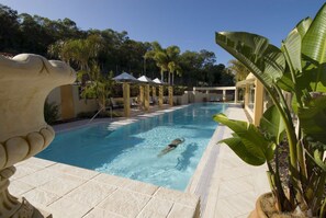 A swimming pool with a person swimming, surrounded by palm trees and covered seating areas. - Noosa Springs Golf Resort & Spa (Noosa Heads)