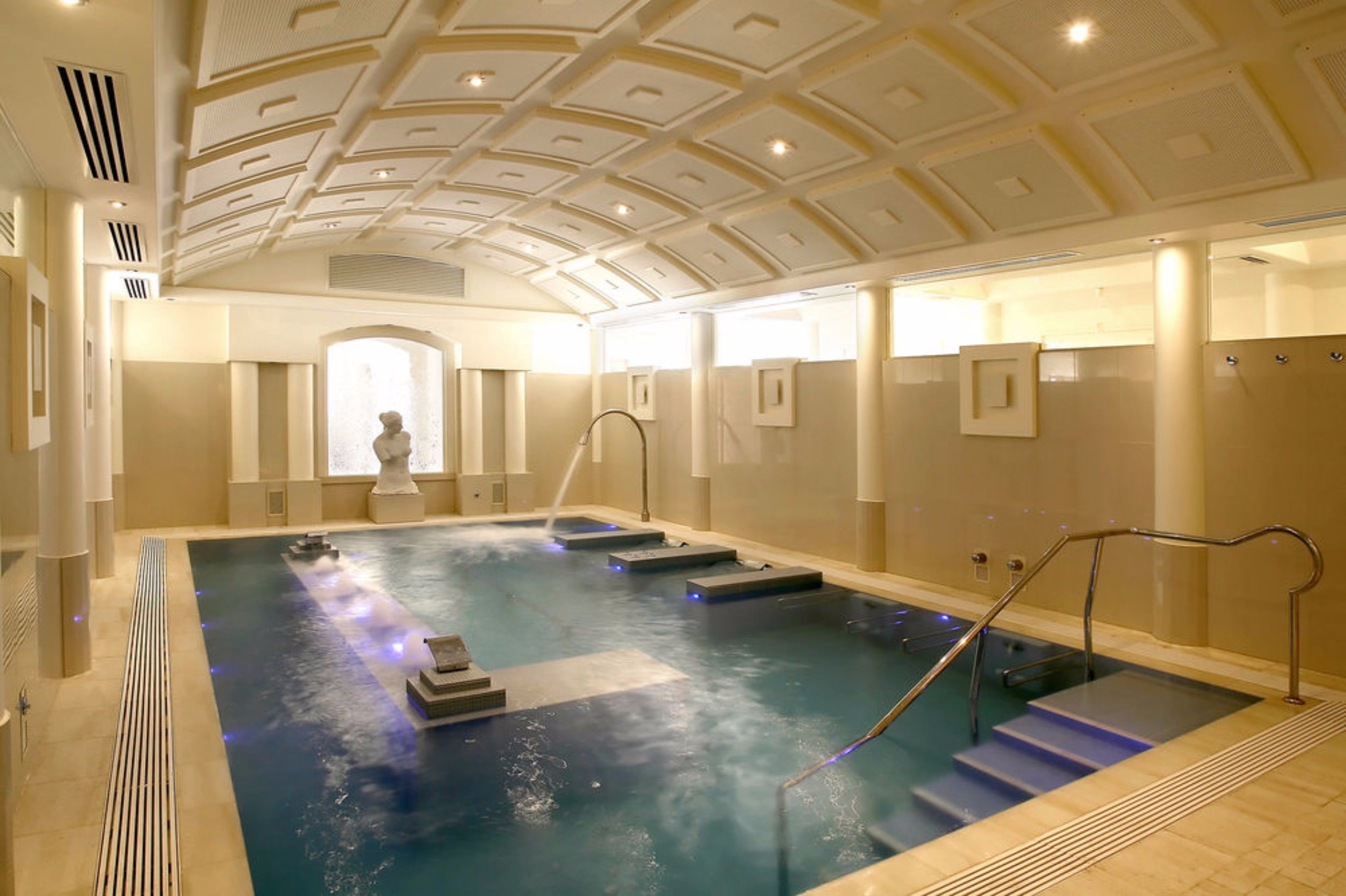 Una amplia piscina interior con escalones que bajan al agua, una estatua al fondo y un techo con diseño geométrico.