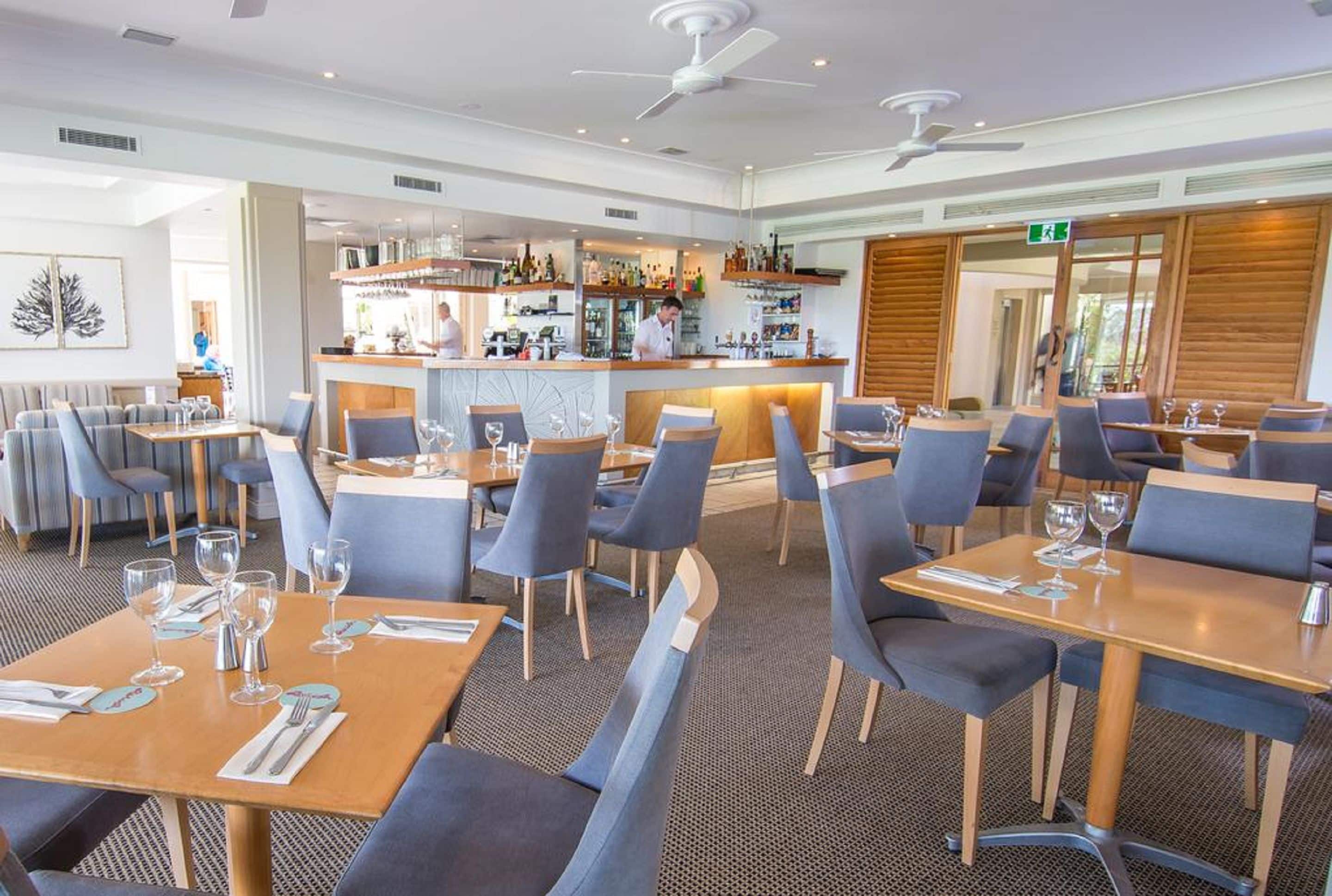 En moderne restaurant med flere spiseborde, stole, et barområde og ventilatorer i loftet.