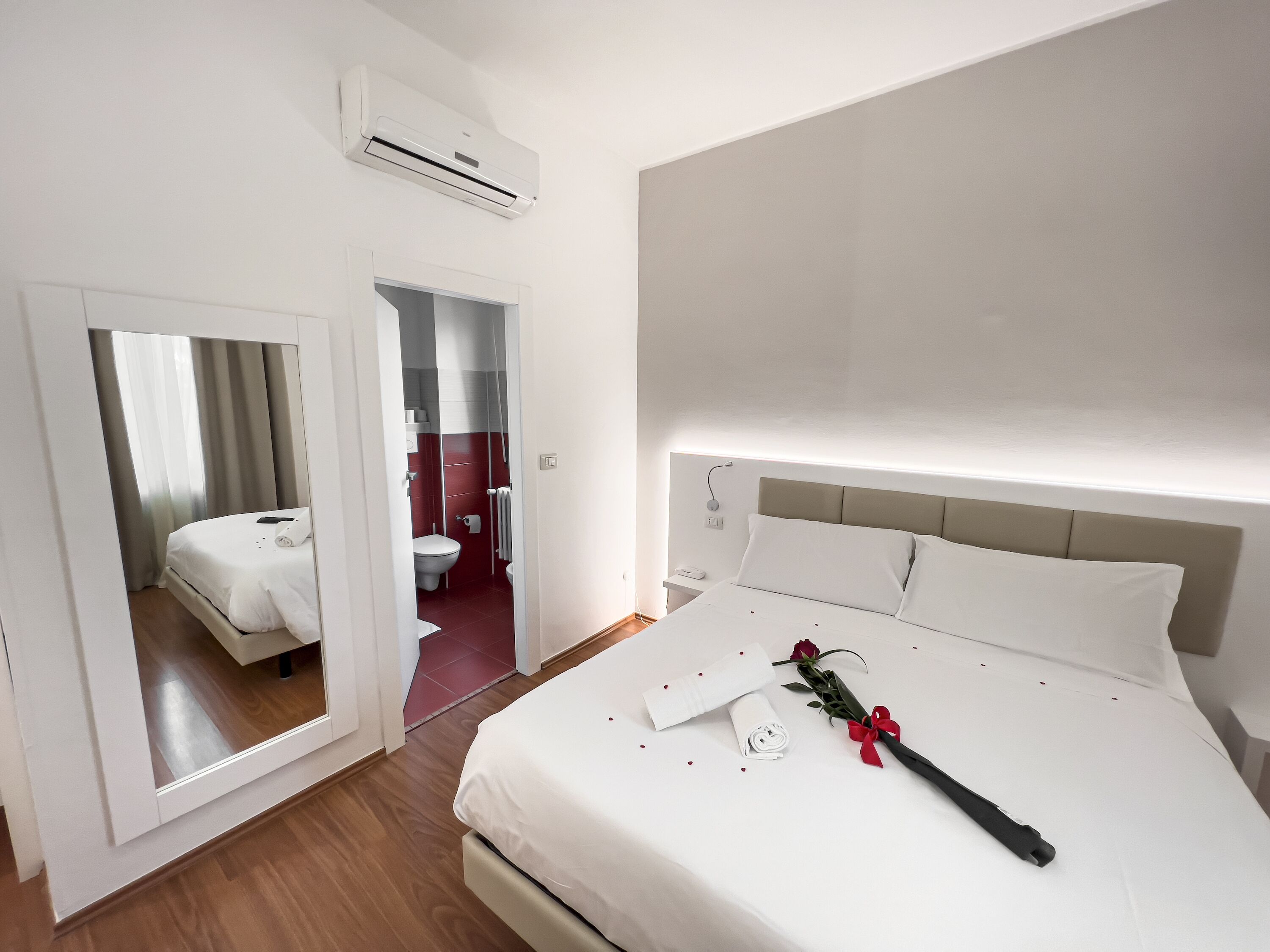 A hotel room with a bed, a bedside table, a nightstand, a mirror, and an air conditioning unit.