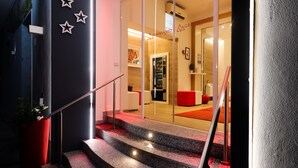 Una entrada de hotel moderna con puerta de vidrio, un banco rojo y una escalera que lleva al interior.