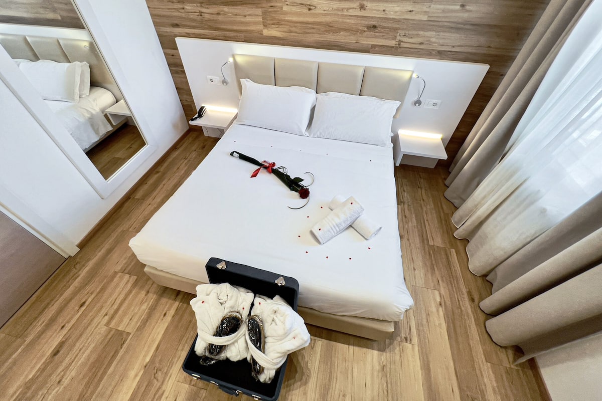 A neatly made bed with white linens, a black suitcase with towels, and a red rose on the bed.