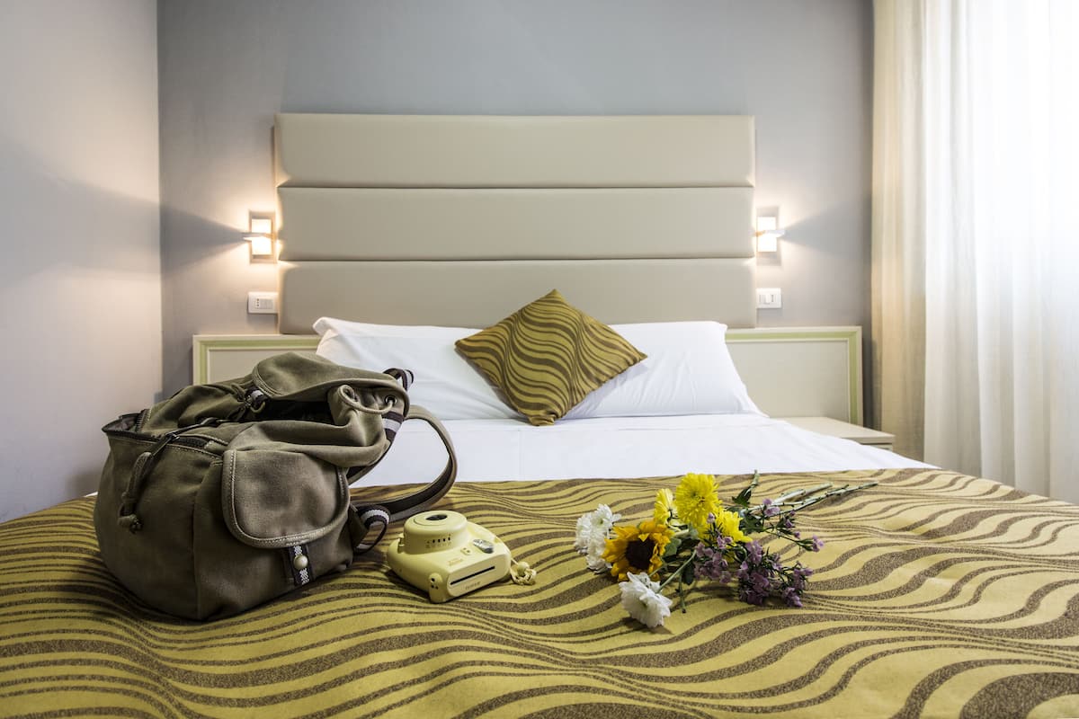 A neatly made bed with a patterned bedspread, a pillow, a bag, a camera, and a bouquet of flowers.