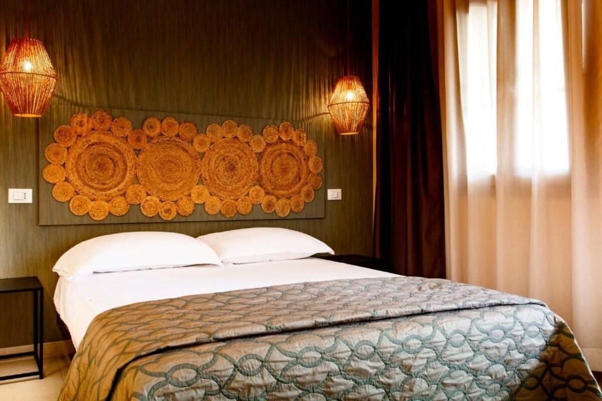 A hotel room with a large bed, a decorative headboard, two wall-mounted lamps, and a window with curtains.