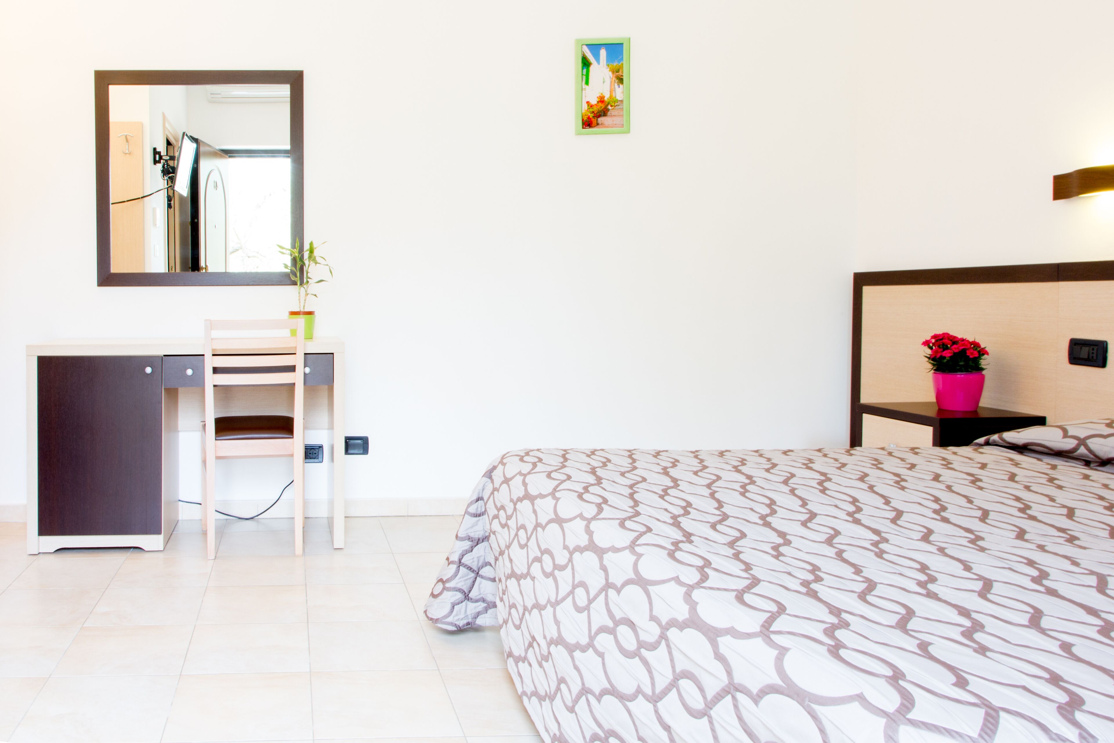 A bedroom with a bed, a desk, a chair, a mirror, a nightstand, and a small potted plant.