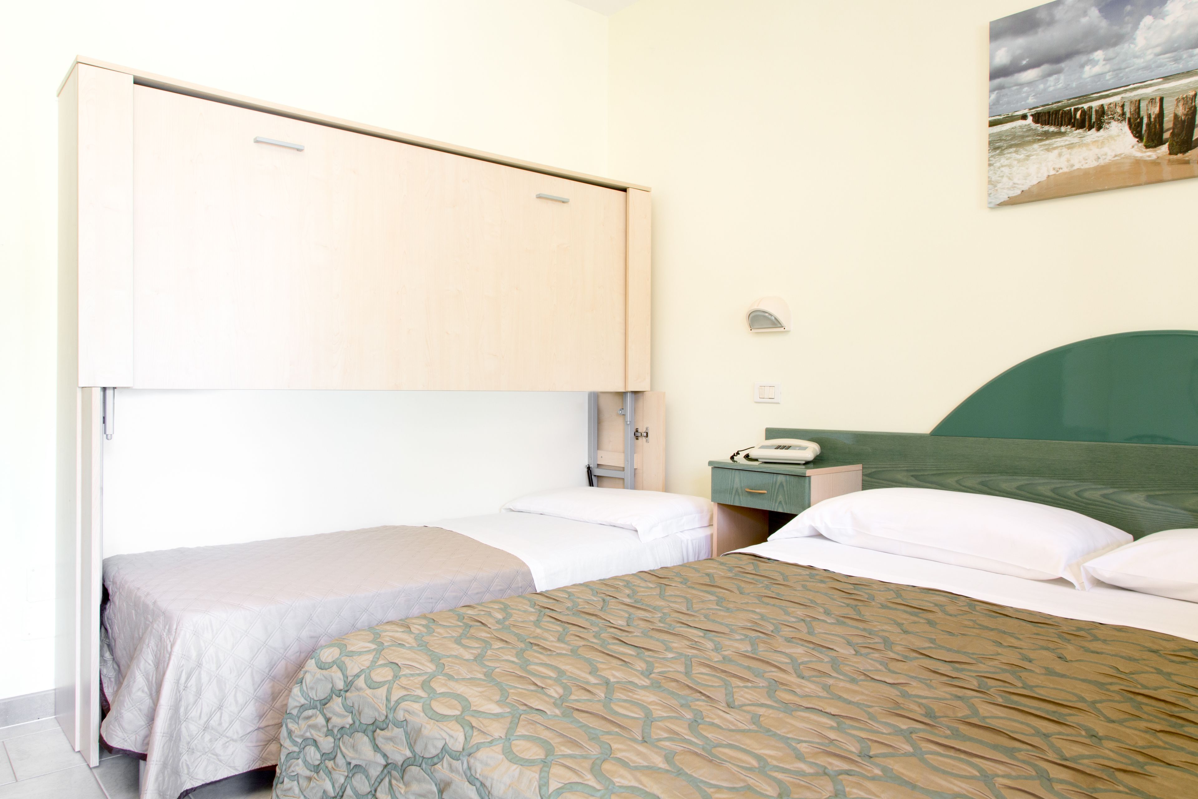 A room with two beds, a wooden wardrobe, a bedside table with a lamp, and a painting on the wall.