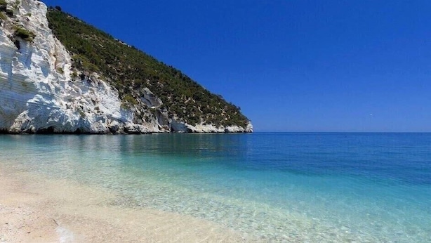 Una playa costera con agua turquesa cristalina, arena blanca y un acantilado rocoso con vegetación.