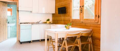 A wooden cabin with a kitchen area featuring white cabinets, a dining table with chairs, a small sink, and a window with a view of greenery.