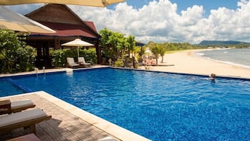 Piscina com espreguiçadeiras e guarda-sóis, vista para a praia e área coberta com cadeiras.