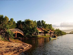 A riverside scene with a wooden bridge, a row of houses with verandas, and a boat on the water.