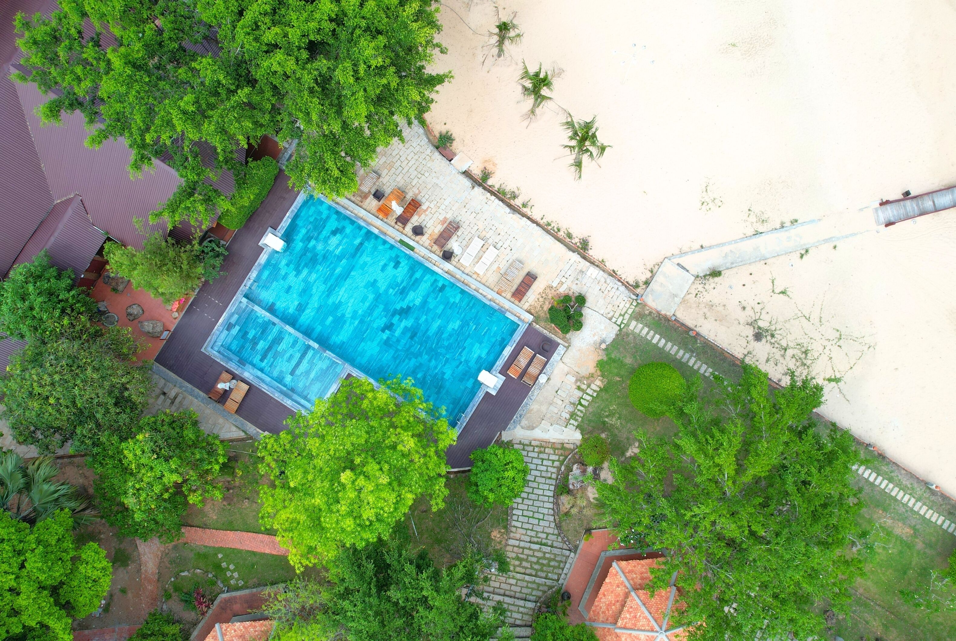 Vue aérienne d’une piscine entourée d’une végétation luxuriante et bordée d’une plage de sable.