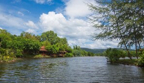 A calm river with a wooden house on the left bank, surrounded by lush greenery and trees.