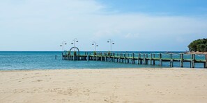 A beach with a wooden pier extending into the sea, featuring a seating area with tables and chairs.