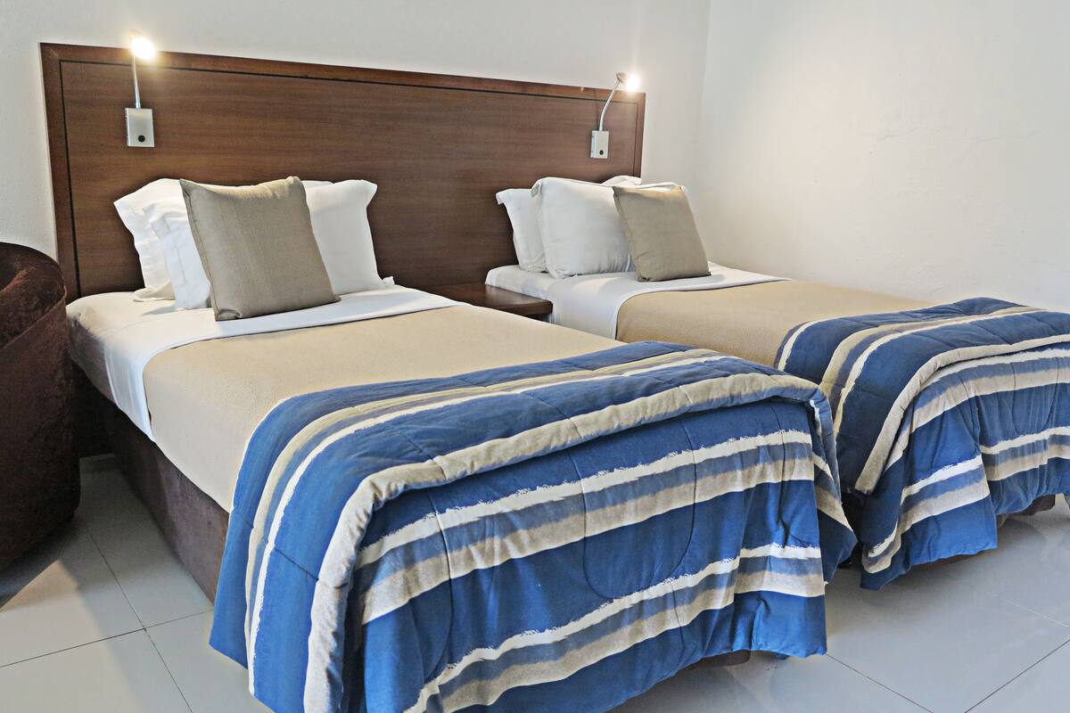 Ein Hotelzimmer mit zwei Betten, jeweils mit einem Holzkopfbrett, weißer Bettwäsche und einer blau gestreiften Decke.