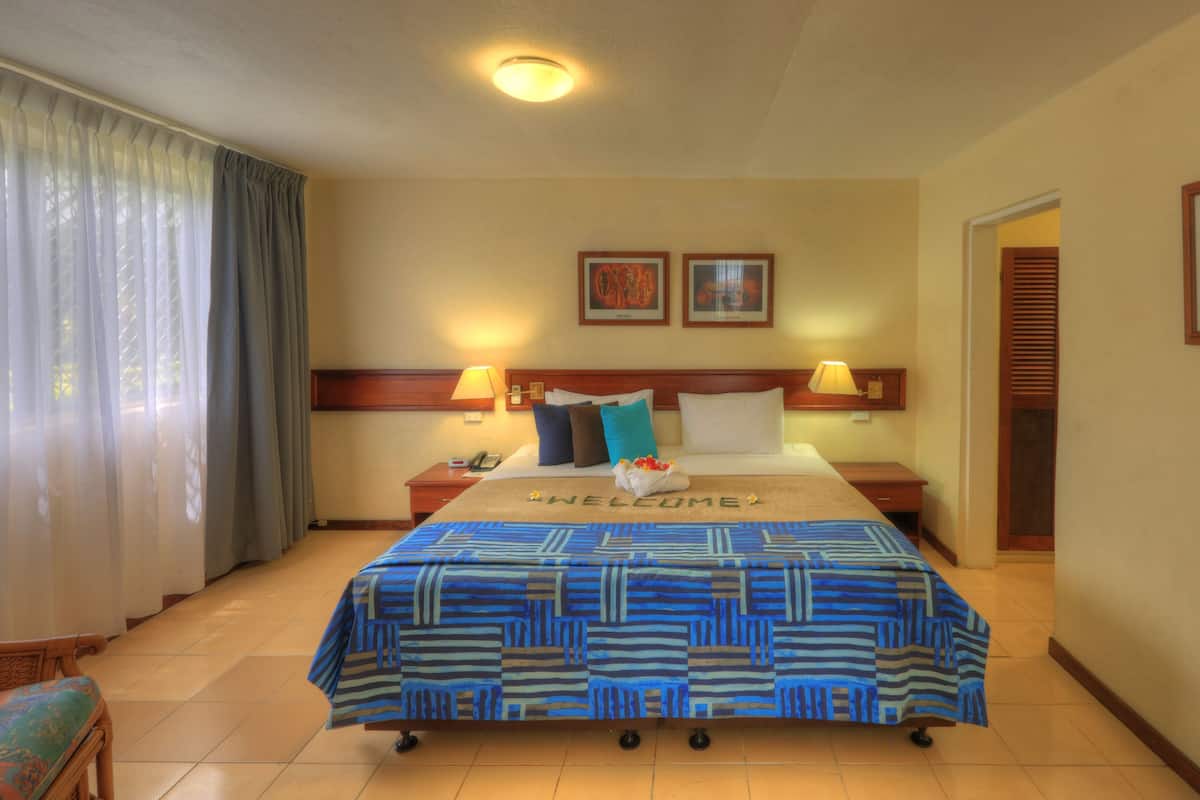 Ein Hotelzimmer mit einem Bett, zwei Nachttischen, einem Sessel, einem Fenster mit Vorhängen und zwei gerahmten Bildern an der Wand.
