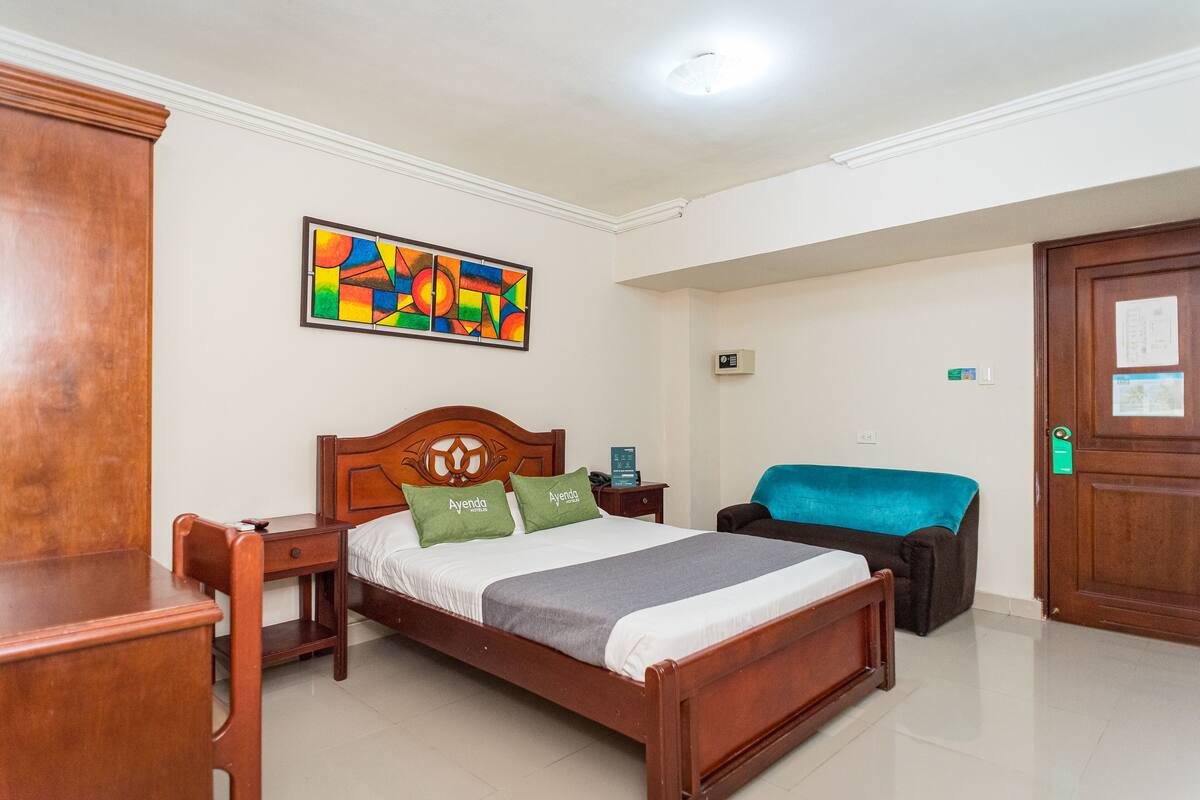 Habitación de hotel con cama de madera, un escritorio y una silla. En la pared, un cuadro abstracto y colorido, y una silla azul en la esquina.