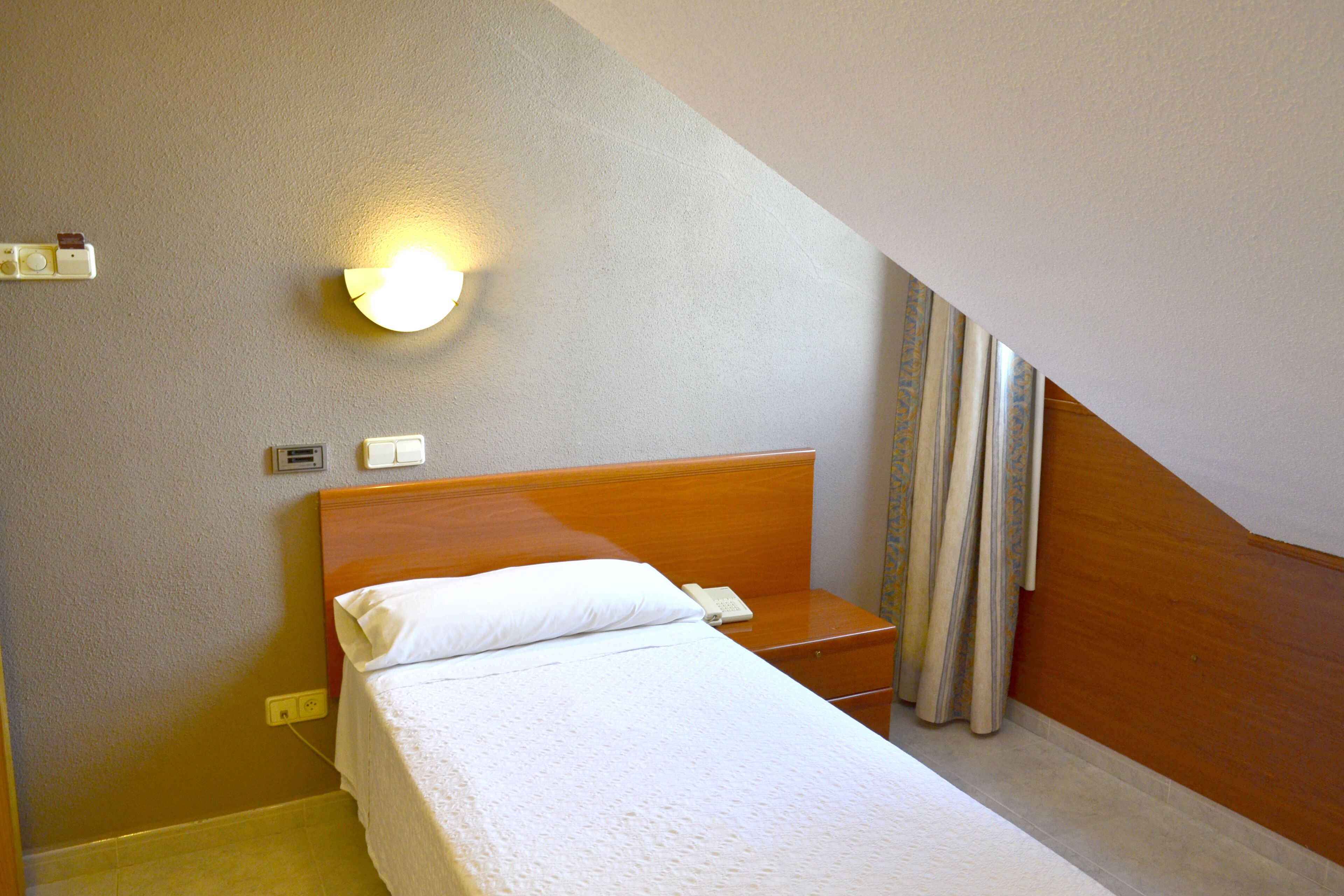 Une chambre d’hôtel avec un lit simple, une table de chevet, une lampe et un rideau.