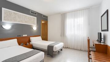 Ein Hotelzimmer mit zwei Betten, einem Schreibtisch mit Computer, einem Fernseher und einem großen Fenster mit Vorhängen.